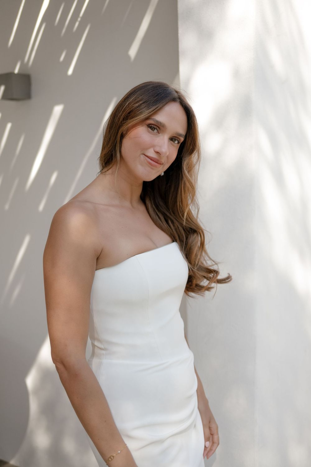 A woman in a white one-shoulder top poses against a bright white wall with dramatic lighting.