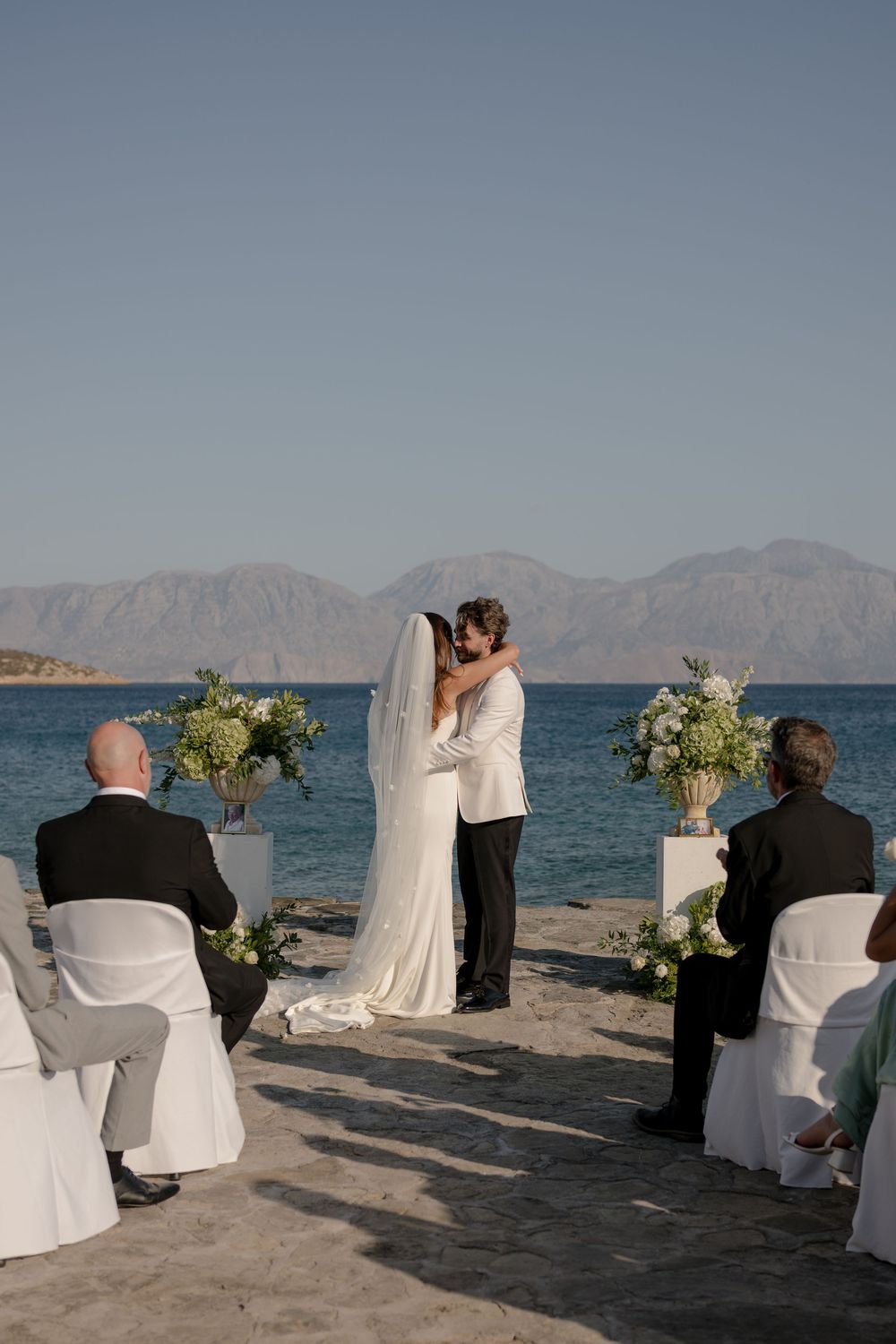 Outdoor beach wedding ceremony setup with white chairs and floral arrangements against a mountainous coastal backdrop.