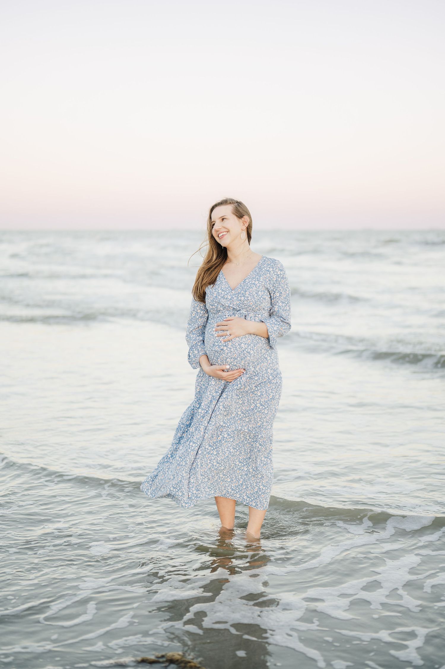 A pregnant woman in a blue floral dress stands in shallow ocean waves during sunset in galveston for a maternity photoshoot