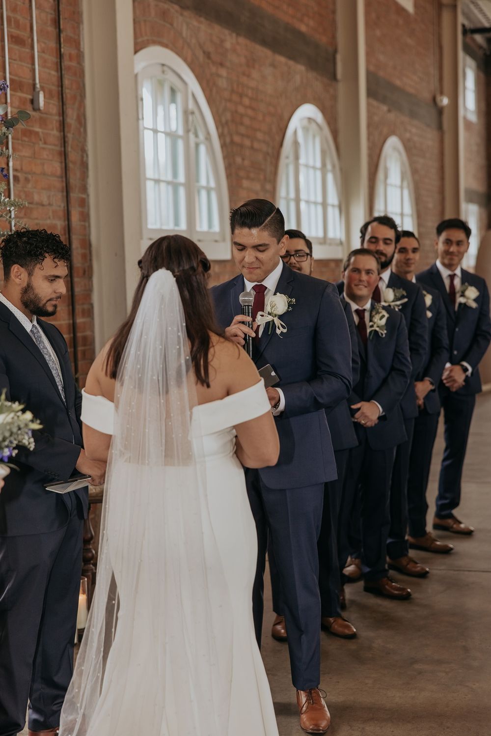 Wedding ceremony in progress along brick corridor with elegant arched windows as attendants look on.