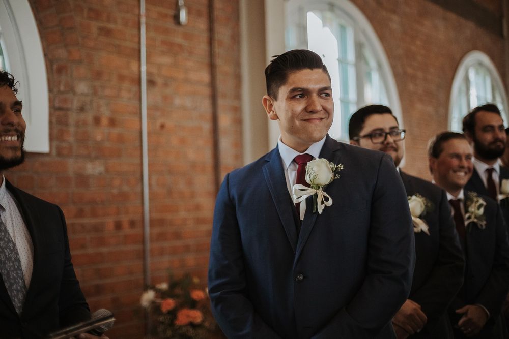 A groom in a navy suit waits for his bride at a wedding venue with exposed brick walls.