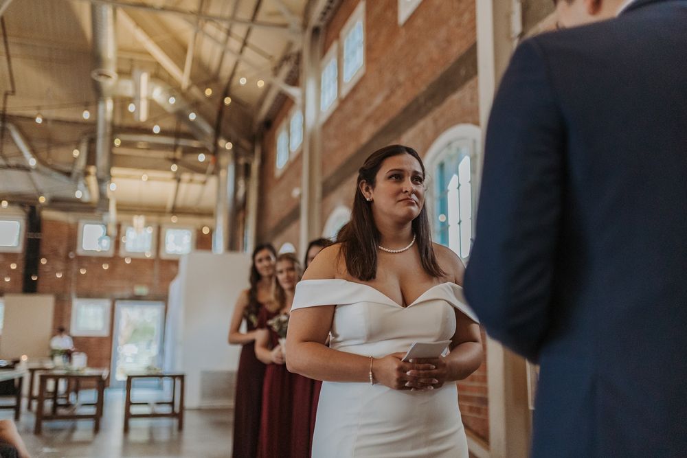 A bride in an elegant off-shoulder white gown stands in a rustic venue with exposed brick walls and string lights.