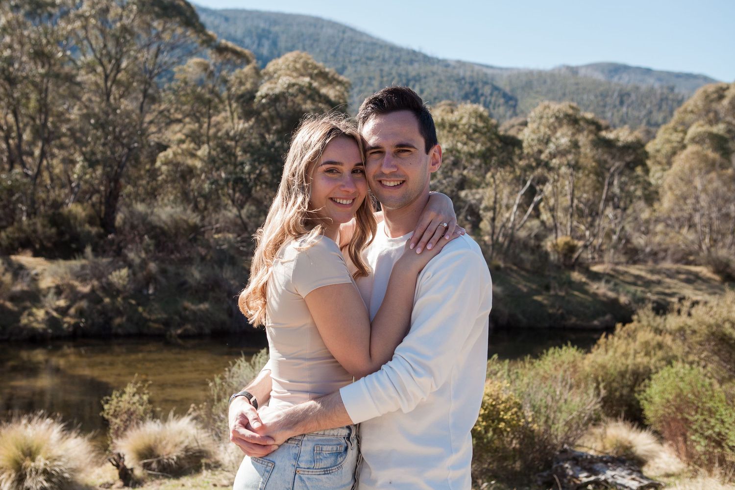 A couple embraces in a romantic outdoor photoshoot against a scenic mountain landscape with natural sunlight.