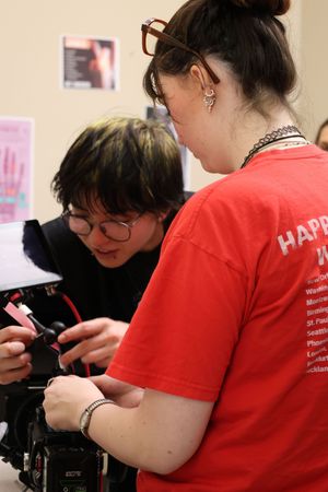 Two people in glasses look down at a device while one wears a red t-shirt that says 'HAPPY' on the back.