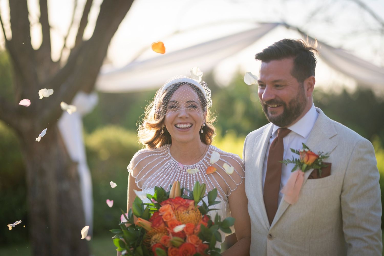 Happy bride and groom laughing outdoors at wedding ceremony with colorful confetti and orange bouquet in golden sunlight.