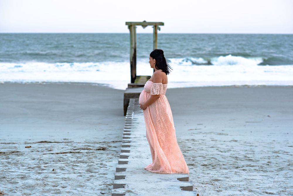 A maternity photoshoot captures a silhouette against the ocean with a wooden pier in the background.