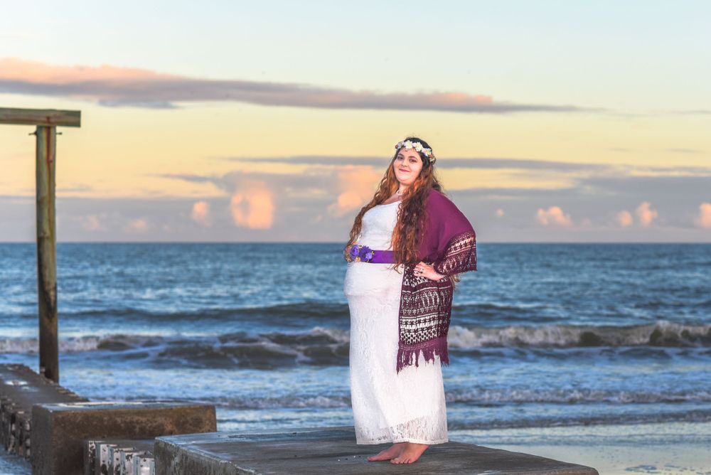A maternity photo session at sunset shows an expectant mother in a white dress and purple shawl by the ocean.