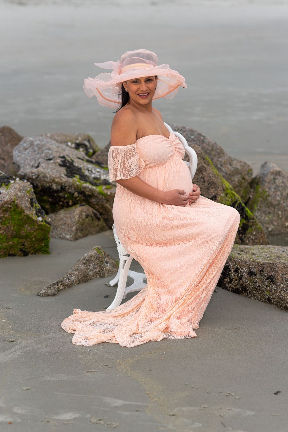 An expectant mother poses in a peach lace gown and sun hat on rocky beach during golden hour.
