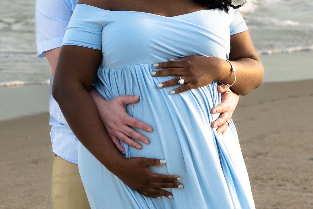 Close up of couple's hands embracing pregnant belly in light blue maternity dress on beach.