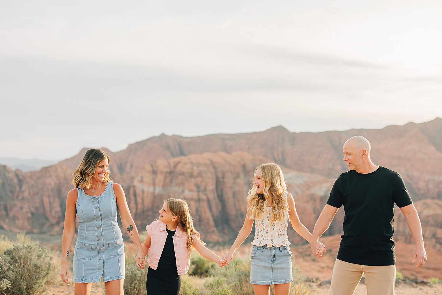Family of four holding hands and laughing in a desert landscape with red rock formations at sunset.