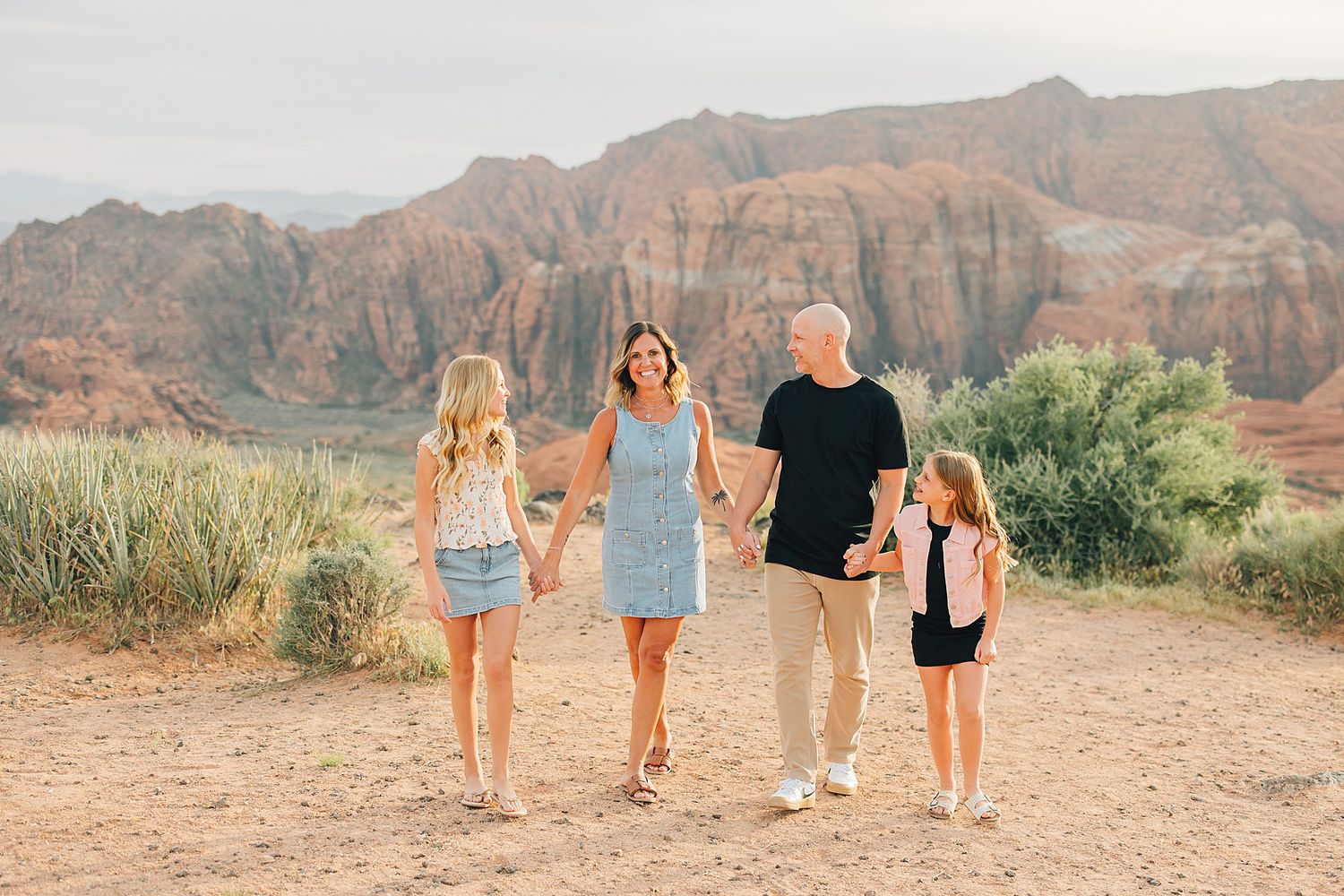 Family of four holding hands walking on red rock desert trail with scenic canyon backdrop.