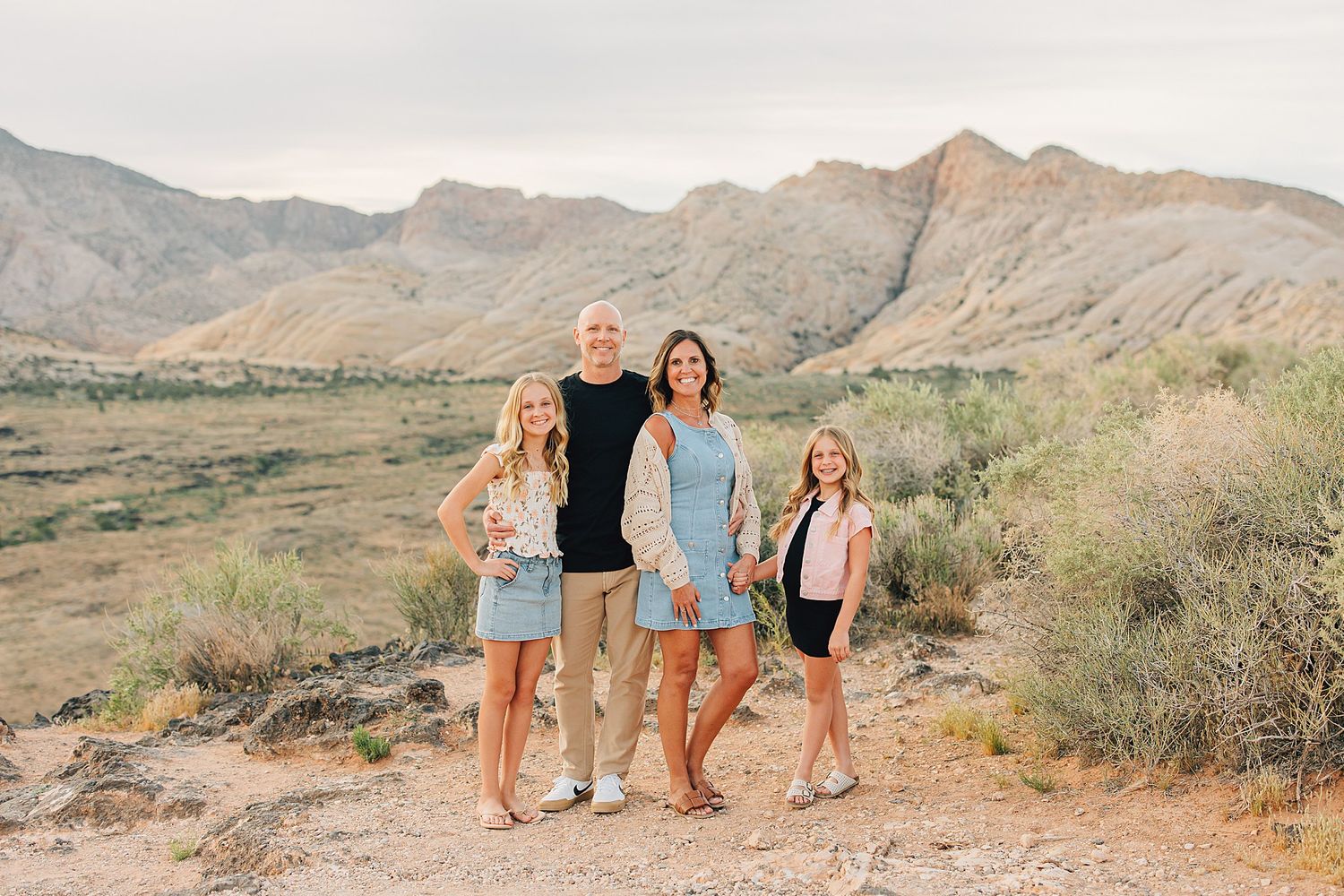 Family of four posing together in a desert landscape with mountains in the background at golden hour.