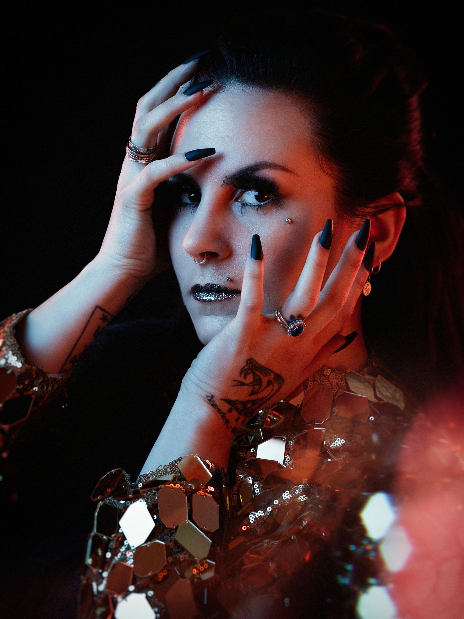 A woman with black nails and gold lips poses dramatically in a sequined gold outfit under moody dark lighting.