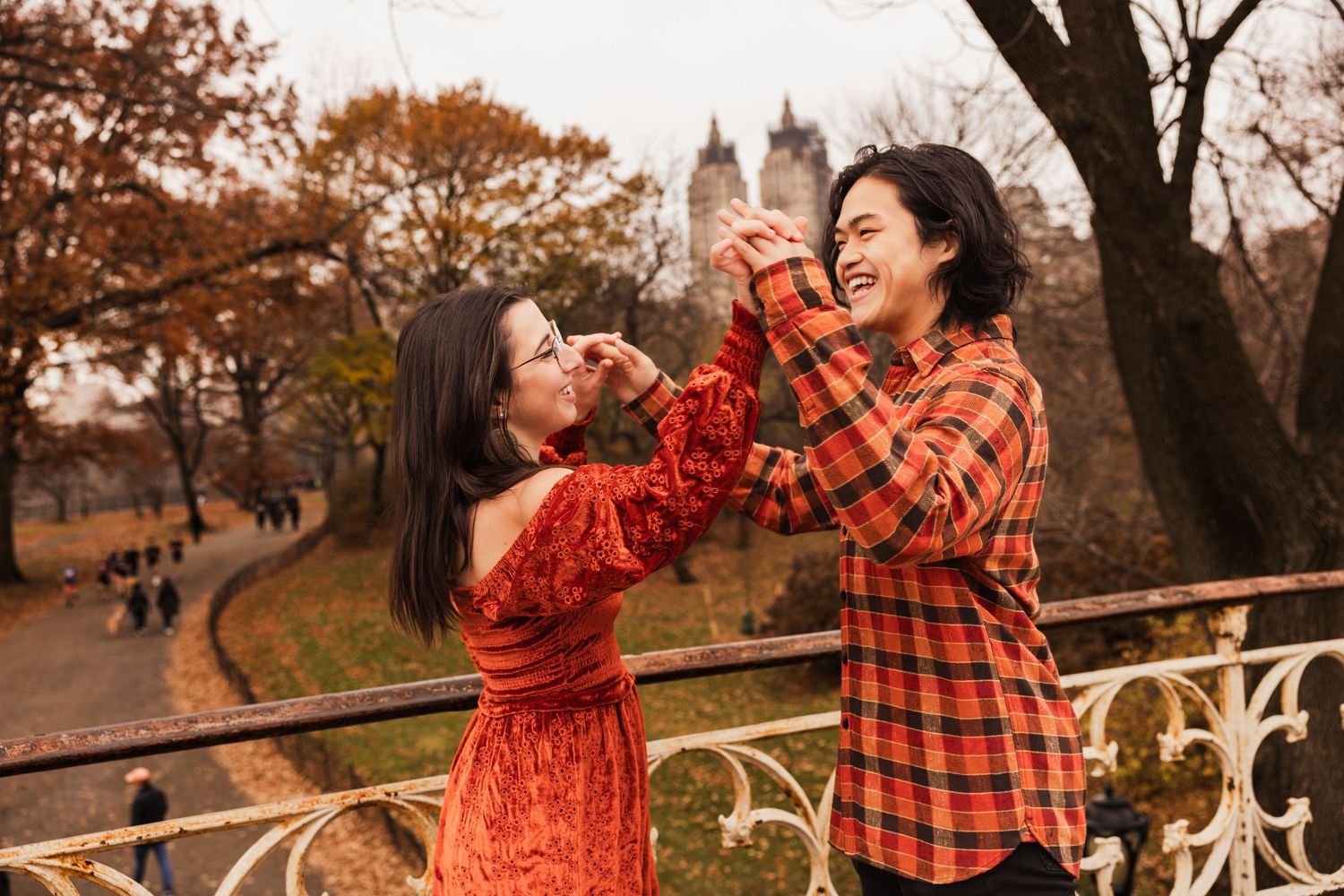 Two people in red and plaid outfits share drinks while posing together on a wooden bridge during autumn.