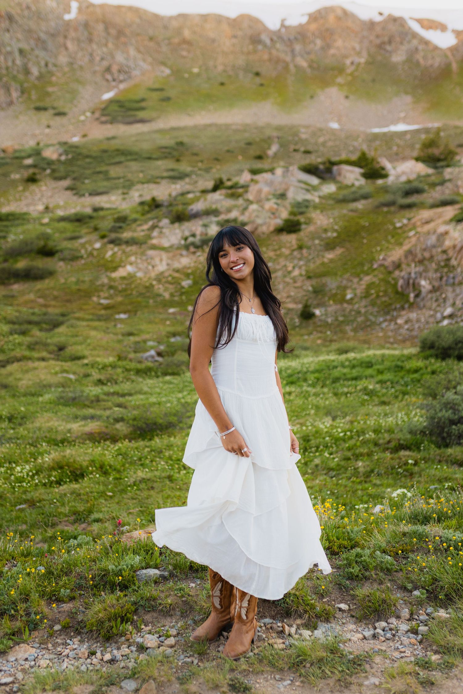A white flowy dress twirls gracefully against a scenic mountain landscape with green meadows and rocky terrain.