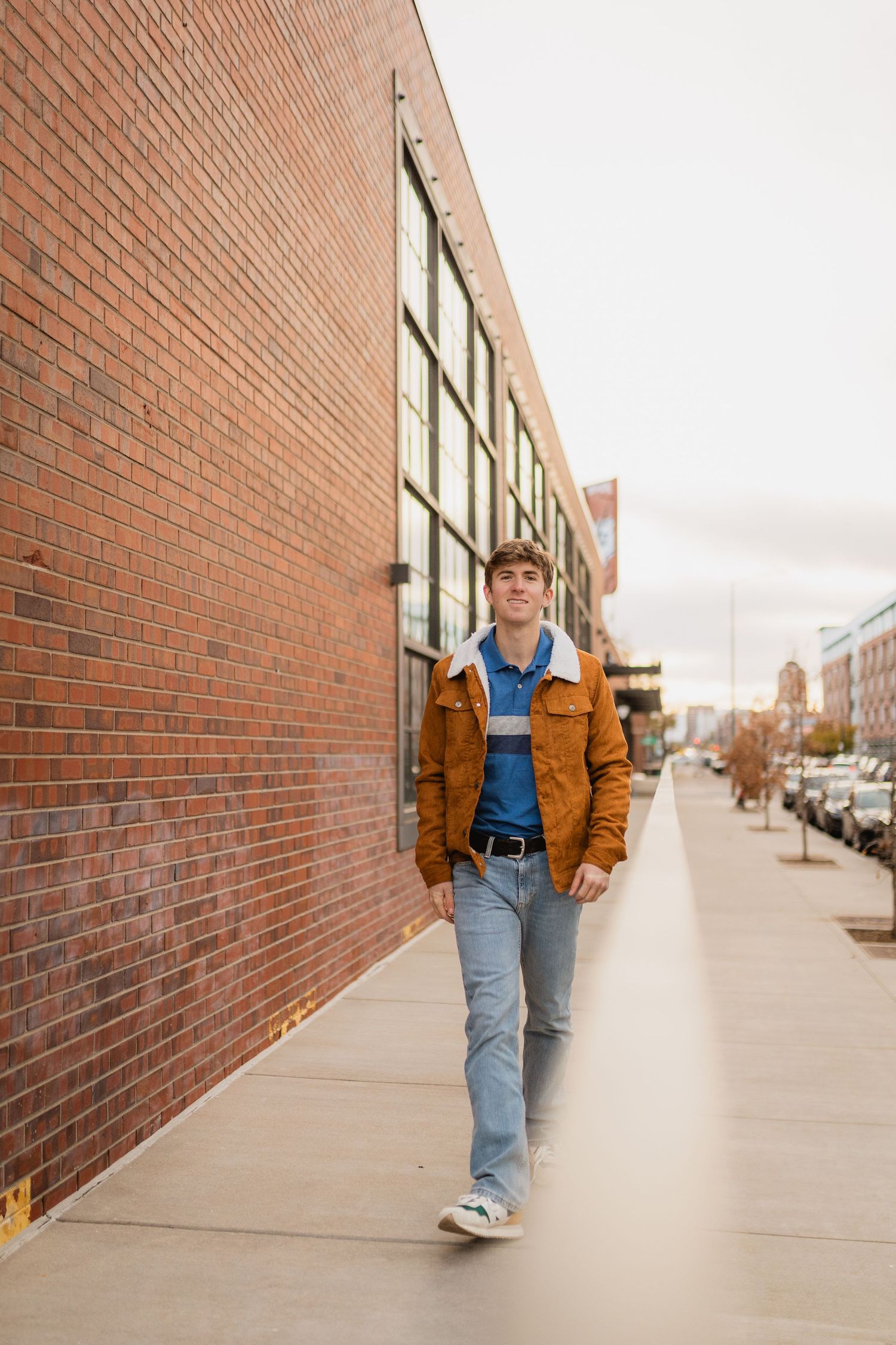 Someone walks along an urban sidewalk wearing a brown jacket and jeans next to a brick building on an autumn day.
