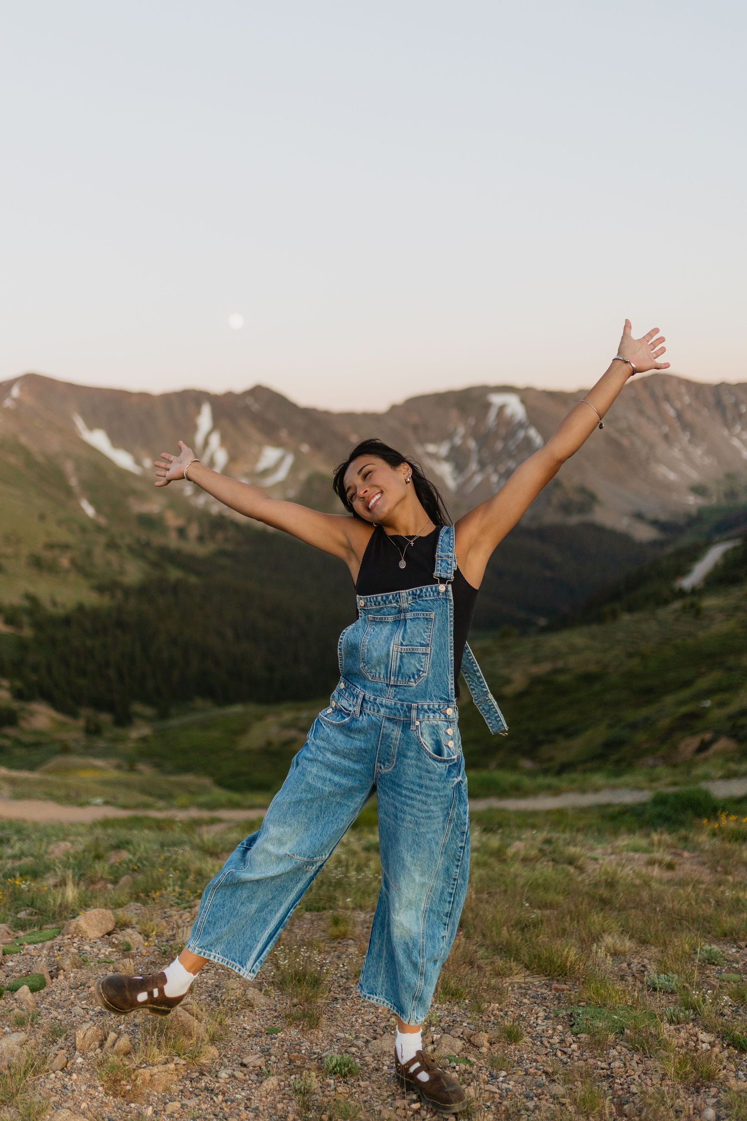 Person in denim overalls dances joyfully against a scenic mountain backdrop at sunset.
