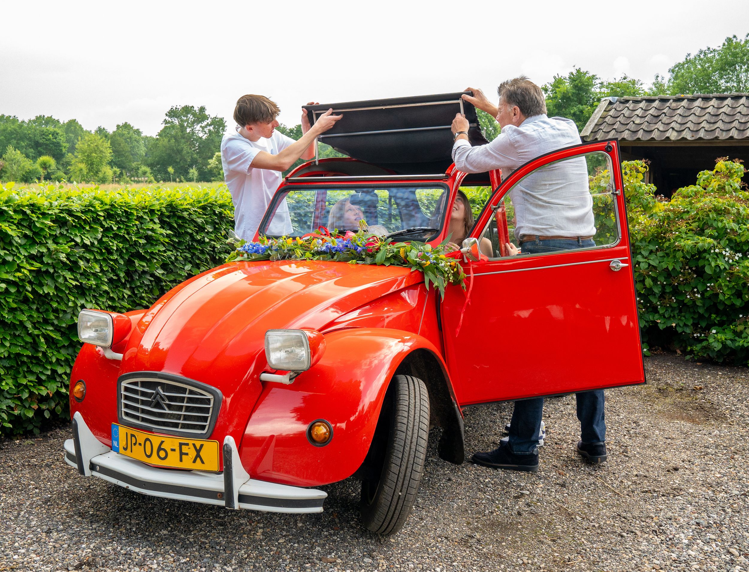 Twee mannen openen het dak van een klassieke rode Citroën 2CV, een trouwauto versierd met bloemen.