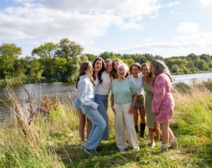 Een groep van zeven vrouwen lacht samen in de buitenlucht, vlakbij een rivier met weelderige groene bomen. familie fotograaf Zwolle