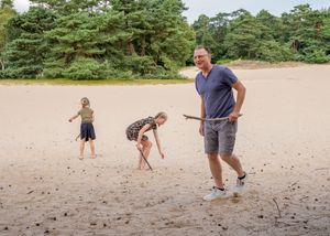 Een man met een stok staat op een zandstrand, met op de achtergrond twee kinderen die bij dennenbomen spelen. familie fotograaf Veluwe