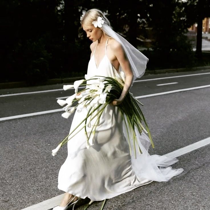 A bride in a flowing white dress walks on a street holding a modern bouquet of long-stemmed flowers and greenery.