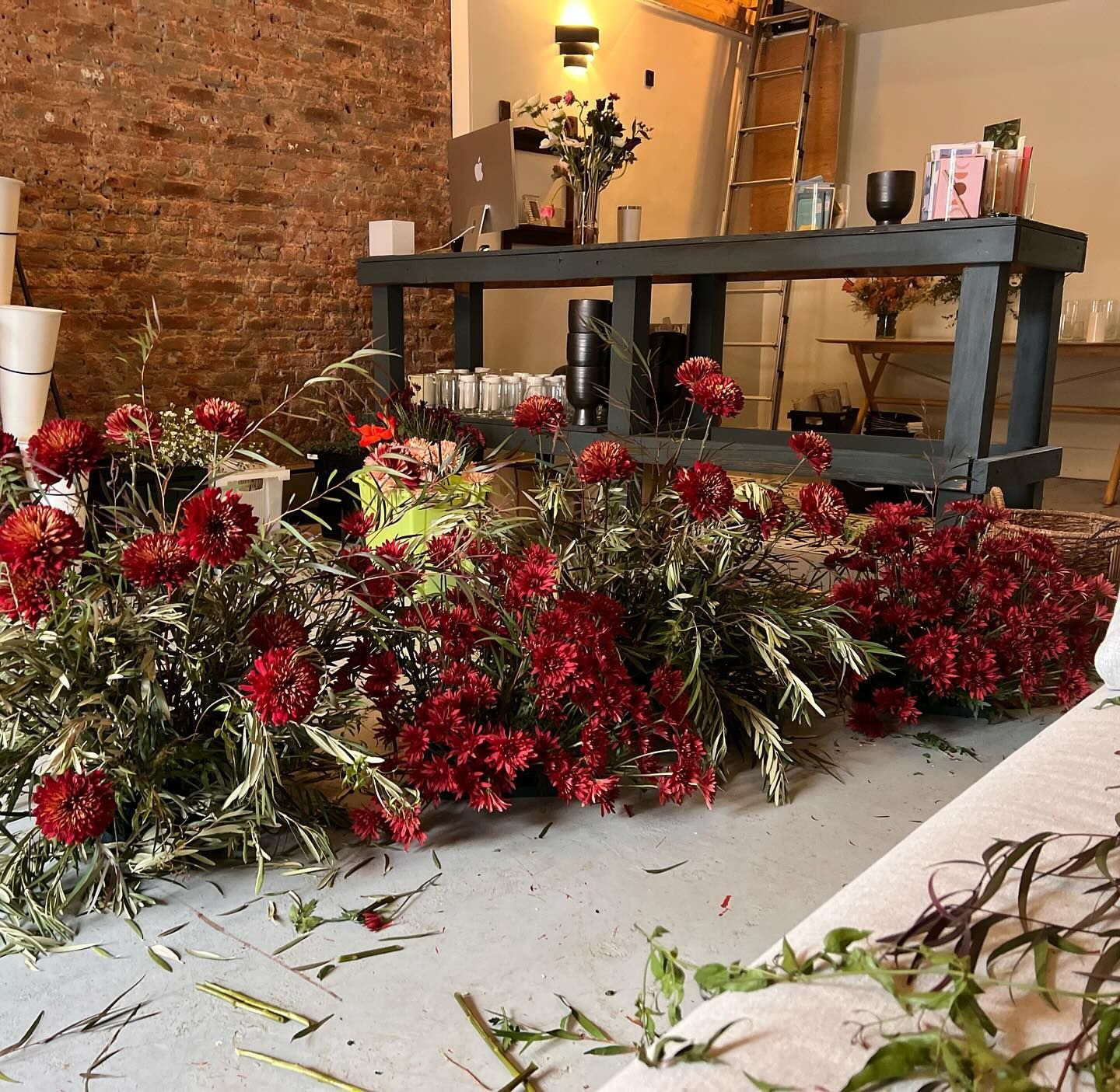 Red floral arrangements and greenery displayed on a white surface against a rustic brick wall with warm lighting.
