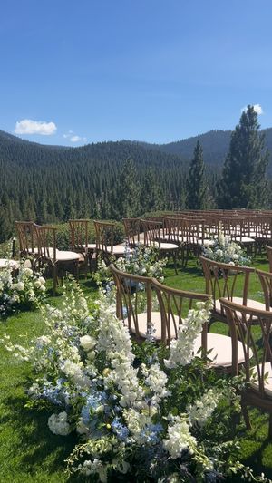 Outdoor mountain wedding ceremony setup with wooden chairs and white floral arrangements against a backdrop of evergreen forests.