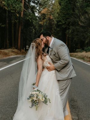 A romantic wedding couple shares an intimate kiss on a tree-lined road while holding a delicate floral bouquet.