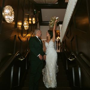 A couple in wedding attire hold hands while ascending a dark staircase illuminated by elegant pendant lights.