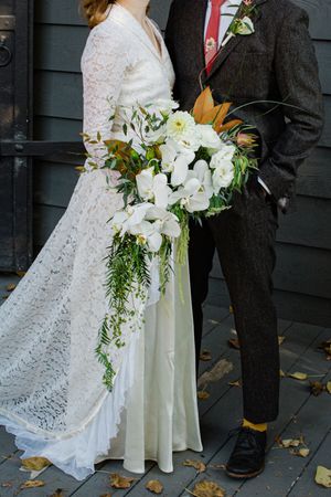 A stunning white lace wedding dress and cascading bouquet of white orchids and greenery stand next to a dark suit.