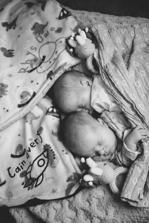 Black and white photo of two sleeping newborn twins wrapped in personalized blankets with crochet toys.