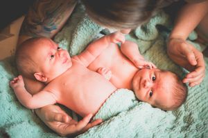Two newborn twins lying side by side on a mint green blanket, with a tattooed parent gently touching them.
