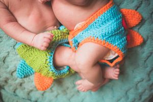 Two newborns in matching crochet dinosaur outfits in blue, green, and orange laying on a teal blanket.