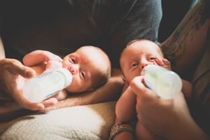 Parent feeding twin newborn babies with milk bottles while cradling them together in arms.
