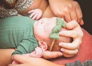 Newborn baby in green outfit and bow headband being cradled by parents hands in a tender moment.