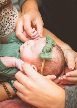 Newborn baby girl in green outfit and bow headband being held by mother and family member.