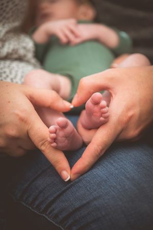 Parent hands forming a heart shape around tiny newborn baby feet, with infant resting in background.