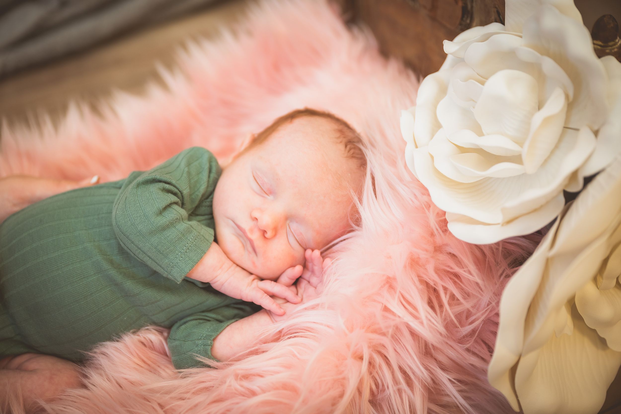 Newborn baby sleeping peacefully on pink fur blanket in green wrap beside white flower decor.