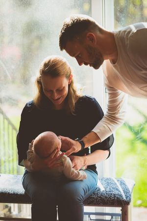 A young couple smiles lovingly at their newborn baby indoors near a large window with natural light.