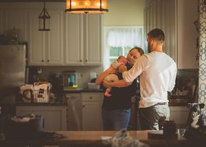 A couple shares a tender moment in a cozy kitchen while one partner gently holds their newborn baby.