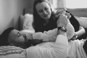 Black and white photo of a bearded father lying down holding newborn baby while mother smiles lovingly nearby.