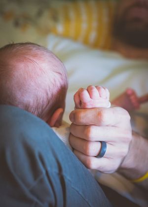 A newborn baby grips a parent's finger, with a wedding ring visible, in a tender bonding moment.