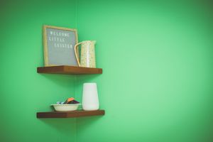 Wooden shelves on a green wall display a welcome sign, a pitcher, and a vase for a nursery decor.