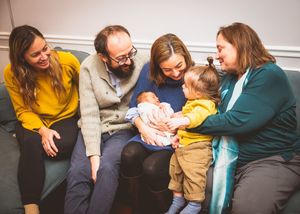 Family group smiling and gathered around a newborn baby and toddler on a cozy sofa indoors.
