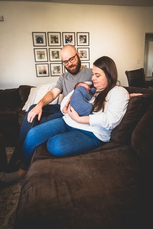 New parents sitting on a brown couch, lovingly gazing at their newborn baby wrapped in a blue blanket.