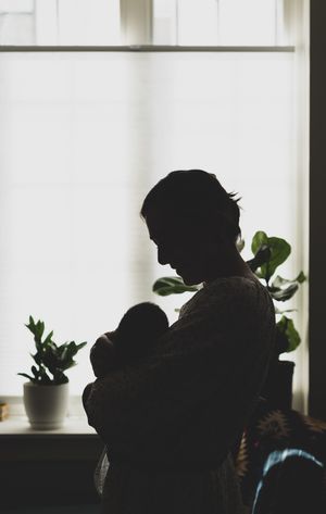 Silhouette of a mother cradling her newborn baby by a bright window with green houseplants in the background.