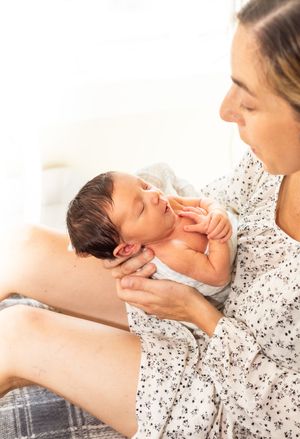 A mother in a floral dress tenderly holds her sleeping newborn baby in her hands, gazing lovingly at her child.