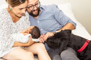 Happy couple sitting on couch introducing their newborn baby to their black dog at home.