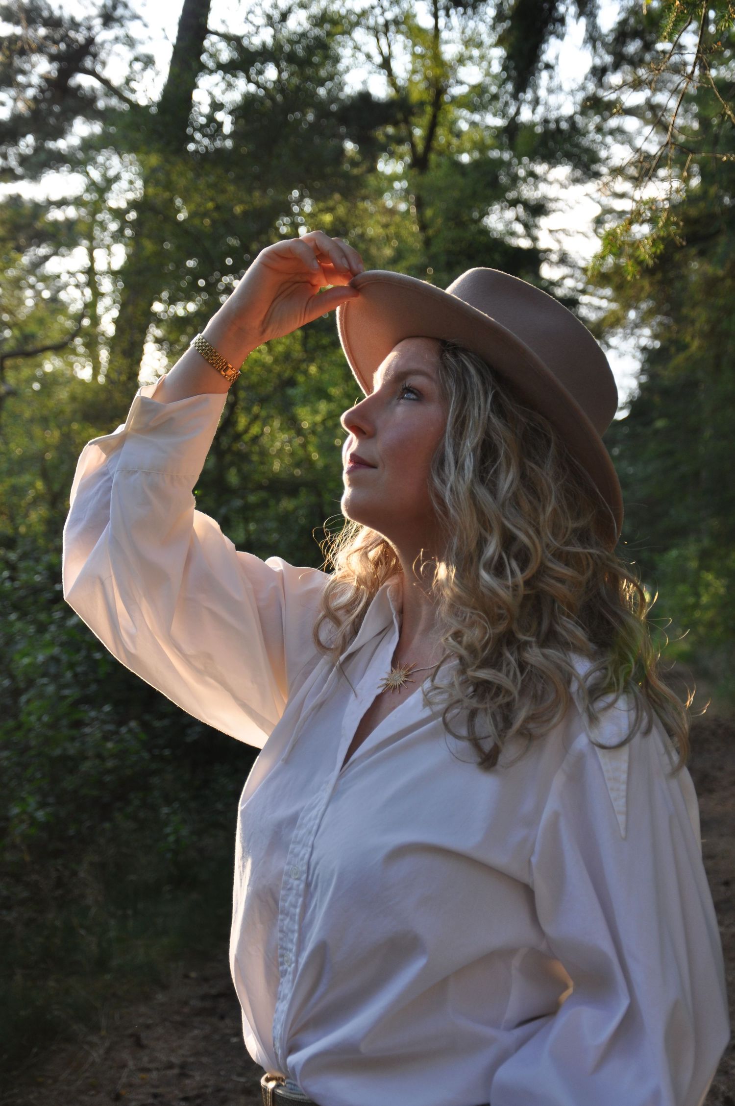 Blonde woman in white blouse and tan cowboy hat posing in sunlit forest.