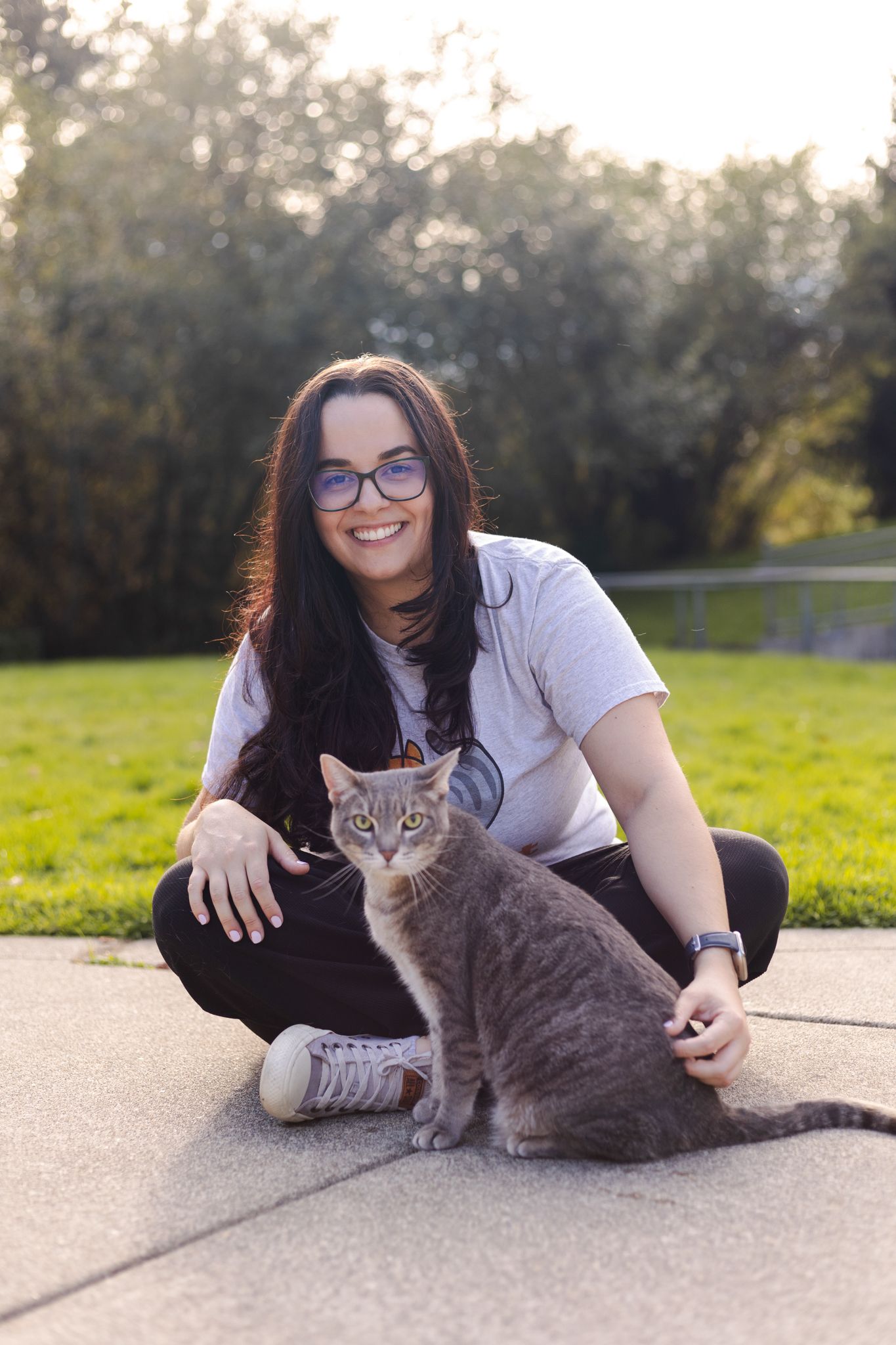 Person in glasses lying on concrete playing with a gray cat outdoors.